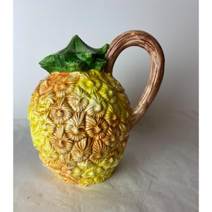 May include: A ceramic pineapple pitcher with a brown handle and green leaves. The pitcher is yellow, orange, and white, with a textured surface. The handle is curved and the leaves are pointed.