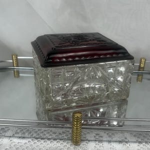 Könnte beinhalten: Eine klare, quadratische Glas-Schmuckschatulle mit einem dunkelroten Deckel. Die Schatulle hat ein geschliffenes Glasdesign, und der Deckel ist mit einem erhabenen Blumenmuster verziert. Die Schatulle steht auf einem Spiegel Tablett mit goldenen Akzenten.