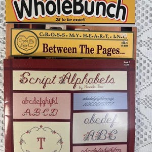 May include: A stack of three cross stitch books. The top book is a yellow magazine titled "Whole Bunch" with the text "25 to be exact!" The middle book is titled "Between The Pages" with the text "C-R-O-S-S. M.Y. H-E-A-R-T,. I.N.C." The bottom book is titled "Script Alphabets" with the text "by Harriette Tew".