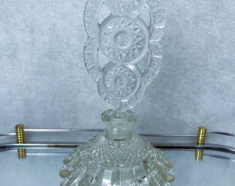 Può includere: Un flacone di profumo in vetro trasparente con un tappo decorativo. La bottiglia ha una base rotonda e pieghettata e un tappo dettagliato con un motivo a stella. La bottiglia è su una superficie a specchio.