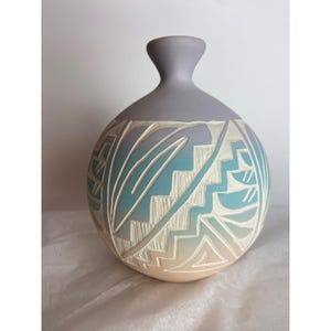 May include: A handcrafted ceramic vase with a round body and a slender neck. The vase features a gradient of colors, transitioning from beige to turquoise and lavender. Intricate white carved patterns adorn the surface, creating a unique decorative piece.