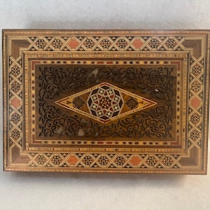 May include: A wooden box with intricate inlaid designs in various shades of brown and red. The box features a geometric pattern on the lid and a carved floral design around the center.