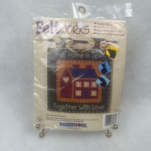May include: A packaged Feltworks craft kit featuring a stitched house design with the text "This Home is Sewn Together With Love." The kit includes iron-on patterns and is 8 x 8 inches.