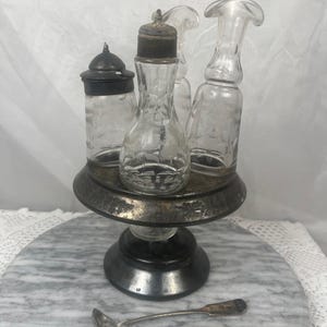 Può includere: Un set di condimenti d'epoca con bottiglie e vasetti di vetro trasparente con coperchi in metallo, appoggiato su un piedistallo argentato. Un piccolo cucchiaio d'argento è davanti. Il set è esposto su una superficie di marmo.