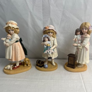 Könnte beinhalten: Drei Porzellanfiguren von Mädchen, die Puppen halten. Jede Figur trägt ein Kleid und einen Hut. Das Mädchen links hält eine Puppe in ihren Armen. Das Mädchen in der Mitte hält eine Puppe in ihrer rechten Hand. Das Mädchen rechts hält eine Puppe in ihrer linken Hand. Jede Figur steht auf einem Sockel mit einem kleinen Accessoire.