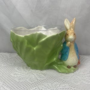 May include: A green ceramic planter shaped like a cabbage with a Peter Rabbit figurine attached. The planter has a clear plastic insert.
