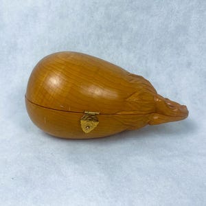 May include: A wooden box with a carved design, featuring a hinged lid and a gold-colored leaf-shaped clasp. The box is a warm, light brown color, with visible wood grain. The box has a rounded shape, with a carved design on one end.