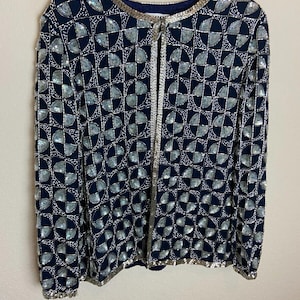 Könnte beinhalten: Marineblaue Jacke mit einem geometrischen Muster aus silbernen Pailletten. Die Jacke hat lange Ärmel und einen runden Ausschnitt, der mit silbernen Pailletten verziert ist. Eine silberne Paillettenborte verläuft entlang der vorderen Öffnung.