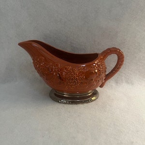 May include: A terracotta coloured ceramic gravy boat with a silver base and a floral design. The boat has a handle on the side.