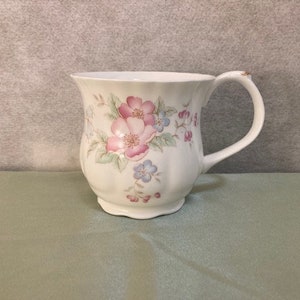 May include: A white ceramic mug with a floral design featuring pink, blue, and green flowers. The mug has a handle and a rounded shape.