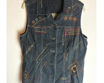 May include: A sleeveless denim vest with colorful embroidered details. The vest features a button-up front, a collar, and various geometric and linear embroidery patterns in shades of red, yellow, blue, and green. The brand label "Chicos" is visible.