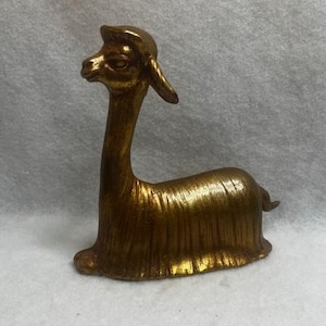 May include: A gold-coloured ceramic llama figurine. The llama is sitting with its head turned to the side. The figurine has a textured surface.