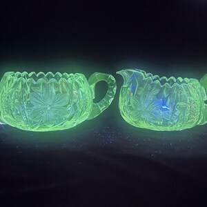 May include: A glowing green glass sugar bowl and creamer set. The set features a floral design and scalloped edges. The handles are curved and the glass has a textured appearance. The set is illuminated against a dark background.
