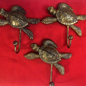 May include: Three gold-colored metal turtle hooks. Each hook is shaped like a turtle with a detailed shell and head. The hooks are mounted on a red background.
