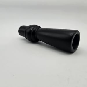 May include: A black bottle stopper with a rounded top and a flared, cylindrical body. The stopper is made of a solid, dark material and is designed to fit into the opening of a bottle. The object is lying on a white surface.