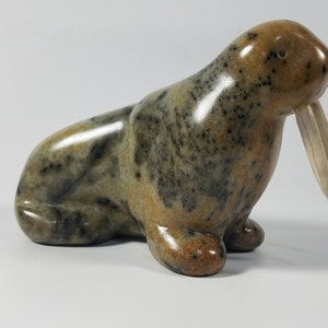 May include: A hand-carved walrus sculpture made of soapstone. The sculpture is brown, green, and black with white tusks. The walrus is sitting with its head turned to the side.