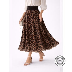 Puede incluir: Falda midi con estampado de leopardo en marrón y negro, con cinturilla negra. La falda se lleva con una blusa blanca y tacones nude. Se sostiene un pequeño bolso de mano blanco. La falda mide unos 76 cm de largo.