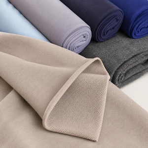 May include: A collection of folded fabric rolls in various colors including beige, light blue, lavender, dark navy, royal blue, and charcoal gray. The fabric features a visible textured weave and is stacked in multiple layers.
