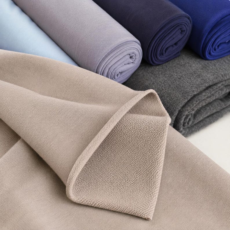 May include: A collection of folded fabric rolls in various colors including beige, light blue, lavender, dark navy, royal blue, and charcoal gray. The fabric features a visible textured weave and is stacked in multiple layers.