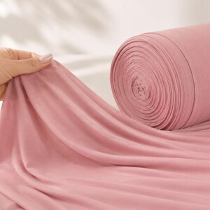 May include: A large roll of solid dusty rose coloured fabric made of a soft, lightweight material. The fabric is draped outward from the cylindrical roll, showing a smooth, matte texture and consistent colour throughout the entire length of the textile.