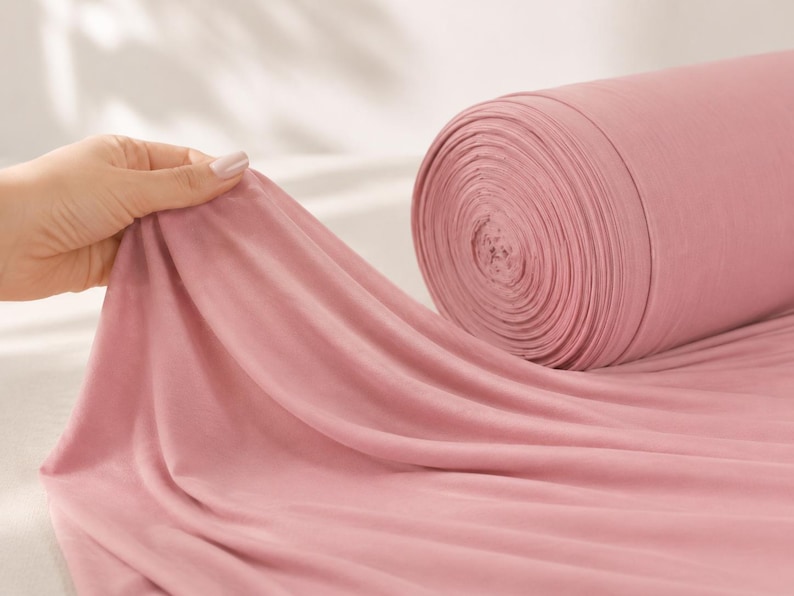 May include: A large roll of solid dusty rose coloured fabric made of a soft, lightweight material. The fabric is draped outward from the cylindrical roll, showing a smooth, matte texture and consistent colour throughout the entire length of the textile.
