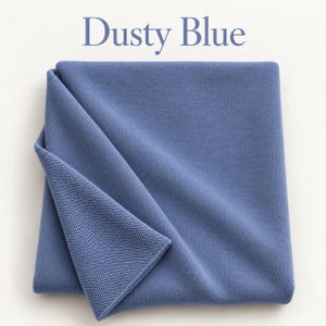 May include: A folded dusty blue knit blanket with a fine, uniform texture. The fabric is a muted, medium-toned blue color. The text "Dusty Blue" is printed in a dark blue serif font above the folded textile, centered on the white background.