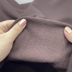 May include: A close-up view of a dark mauve fabric with a visible looped texture on the interior side. The material appears thick and soft, showing a consistent knit pattern across the surface, with the fabric folded to display the inner lining.