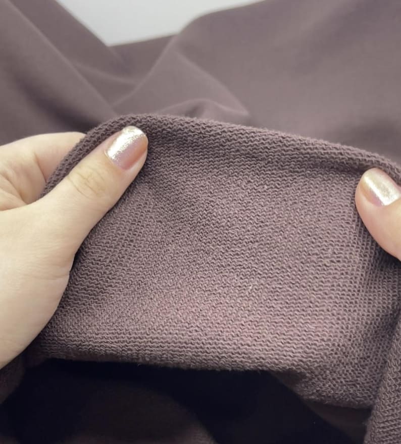 May include: A close-up view of a dark mauve fabric with a visible looped texture on the interior side. The material appears thick and soft, showing a consistent knit pattern across the surface, with the fabric folded to display the inner lining.