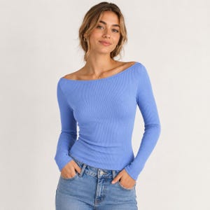 May include: A periwinkle blue long-sleeve top featuring a wide boat neckline and a ribbed knit texture. The garment is fitted through the torso and arms, extending to the waist, and is paired with light blue denim jeans with a visible metal button.