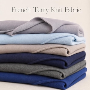 May include: A stack of folded French Terry Knit Fabric in various colors including light gray, pale blue, beige, navy blue, dark gray, and royal blue. The fabric features a smooth top surface and a looped texture on the underside.