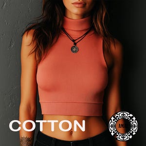 Op de afbeelding: Een mouwloze, geribbelde koraalkleurige crop top met een hoge col. Het kledingstuk wordt gedragen met een zwart koordhalsketting met een ronde hanger. Het woord COTTON staat in witte letters linksonder in de afbeelding gedrukt.