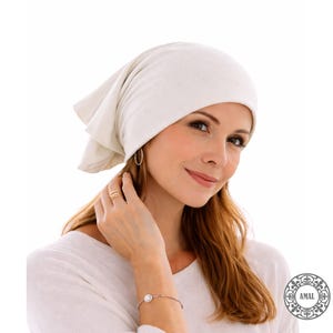 May include: A woman wearing a white headscarf made of soft, draped fabric. She is also wearing gold hoop earrings, a gold ring, and a bracelet with a pearl. The background is white.