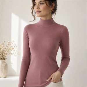 May include: A dusty rose long-sleeve mock neck top featuring a fine ribbed knit texture. The garment has a fitted silhouette with a high neckline that sits close to the throat and full-length sleeves, crafted from a soft, stretchy fabric material.
