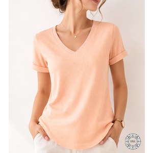 May include: A peach-colored V-neck t-shirt with rolled-up sleeves. The shirt has a ribbed texture and a curved hemline. The model is wearing a gold necklace and watch. The shirt is a casual top.