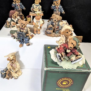 May include: A collection of Boyd's Bears figurines, each depicting a bear in various outfits and poses. Some bears are dressed in patriotic attire, while others are in school uniforms or winter clothing. A green box with the Boyd's Bears logo is also present.