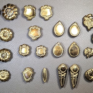 May include: A collection of assorted brass-toned metal buttons featuring various shapes and designs, including floral patterns, shield motifs, oval frames, and decorative embossed details, arranged in rows on a textured gray surface.