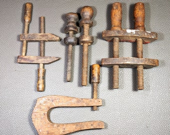 May include: A collection of antique wooden clamps and vises, featuring various sizes and designs. The clamps have wooden handles and screw mechanisms, showing signs of age and use. The tools are arranged on a dark surface.
