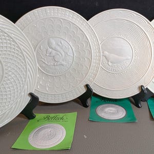 May include: Four cream-coloured decorative plates with embossed designs, including a squirrel, a bird, and a fox. Each plate is displayed on a black stand, with green cards visible below. The plates have intricate patterns and textures.