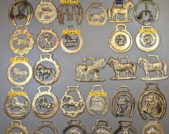 May include: A collection of brass horse brasses featuring various equestrian motifs, including horses, horse heads, and horseshoes. Several pieces are marked with the word SOLD in yellow text. These decorative metal ornaments vary in shape.
