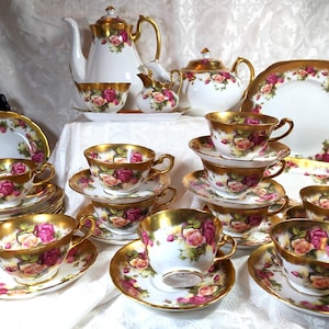 May include: A vintage tea set featuring white porcelain pieces with gold trim and pink rose floral designs. The set includes a teapot, creamer, sugar bowl, cups, saucers, and plates, perfect for a formal tea party or display.