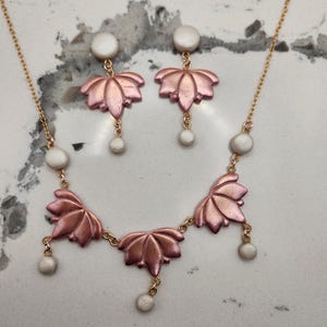 May include: A jewelry set featuring a necklace and earrings. The necklace and earrings have a lotus flower design in a metallic rose gold color. The earrings and necklace have white pearl accents and are on a gold chain.