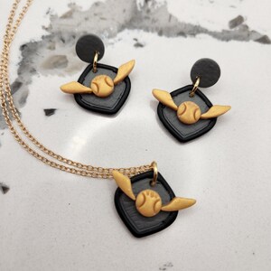 May include: A gold-colored necklace and matching earrings set. The jewelry features a black square pendant with a gold-colored ball and wings design. The earrings have a black stud. The jewelry is displayed on a white and gray marble surface.