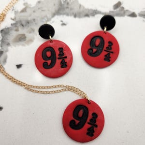 May include: A jewelry set featuring a necklace and earrings. The set includes a gold chain necklace and a pair of earrings, each with a red circular pendant. The pendants display the number "9 3/4" in black.