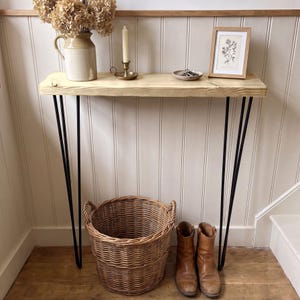 May include: A narrow console table with a light-toned wooden top and four black metal hairpin legs. On the table sits a beige ceramic pitcher with dried flowers, a brass candle holder with a taper candle, a small dish with keys, and a framed botanical print.