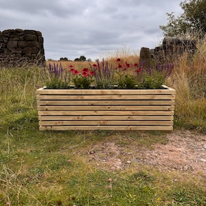 May include: A wooden planter box with a slatted design. The planter is filled with a variety of plants, including purple flowers and green foliage. The planter is sitting on a grassy surface.