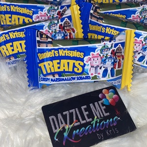 May include: A close-up of a blue and yellow treat bag with the text "Daniel's Krispies Treats" and a picture of cartoon characters. The bag is on a white fluffy surface.