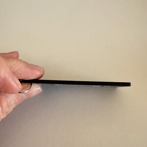 May include: A thin, flat, rectangular black keychain with a smooth edge profile. The object features a small metal ring attached to one side, designed for holding keys. The side view shows the slim, uniform thickness of the black material.