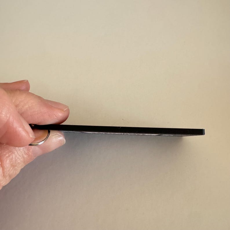 May include: A thin, flat, rectangular black keychain with a smooth edge profile. The object features a small metal ring attached to one side, designed for holding keys. The side view shows the slim, uniform thickness of the black material.