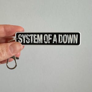 Peut inclure: Un porte-clés rectangulaire noir avec une inscription blanche vieillie qui indique SYSTEM OF A DOWN. Le porte-clés est doté d'un anneau fendu en métal argenté fixé à une extrémité par une petite boucle métallique pour attacher les clés.