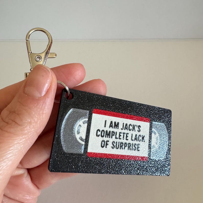 May include: A black rectangular keychain shaped like a VHS tape with a textured surface. The center label is white with a red border and features the black text: I AM JACK'S COMPLETE LACK OF SURPRISE. It includes a silver metal lobster claw clasp.